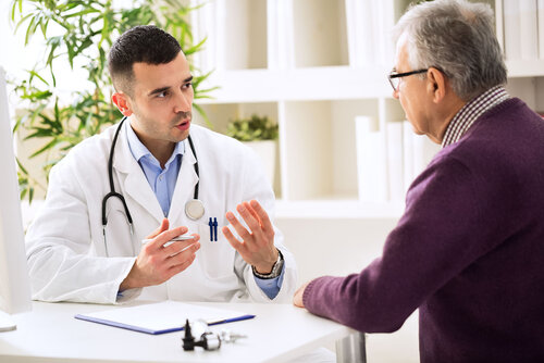 Ein Arzt und ein älterer Patient unterhalten sich an einem Tisch im Sprechzimmer einer Praxis.